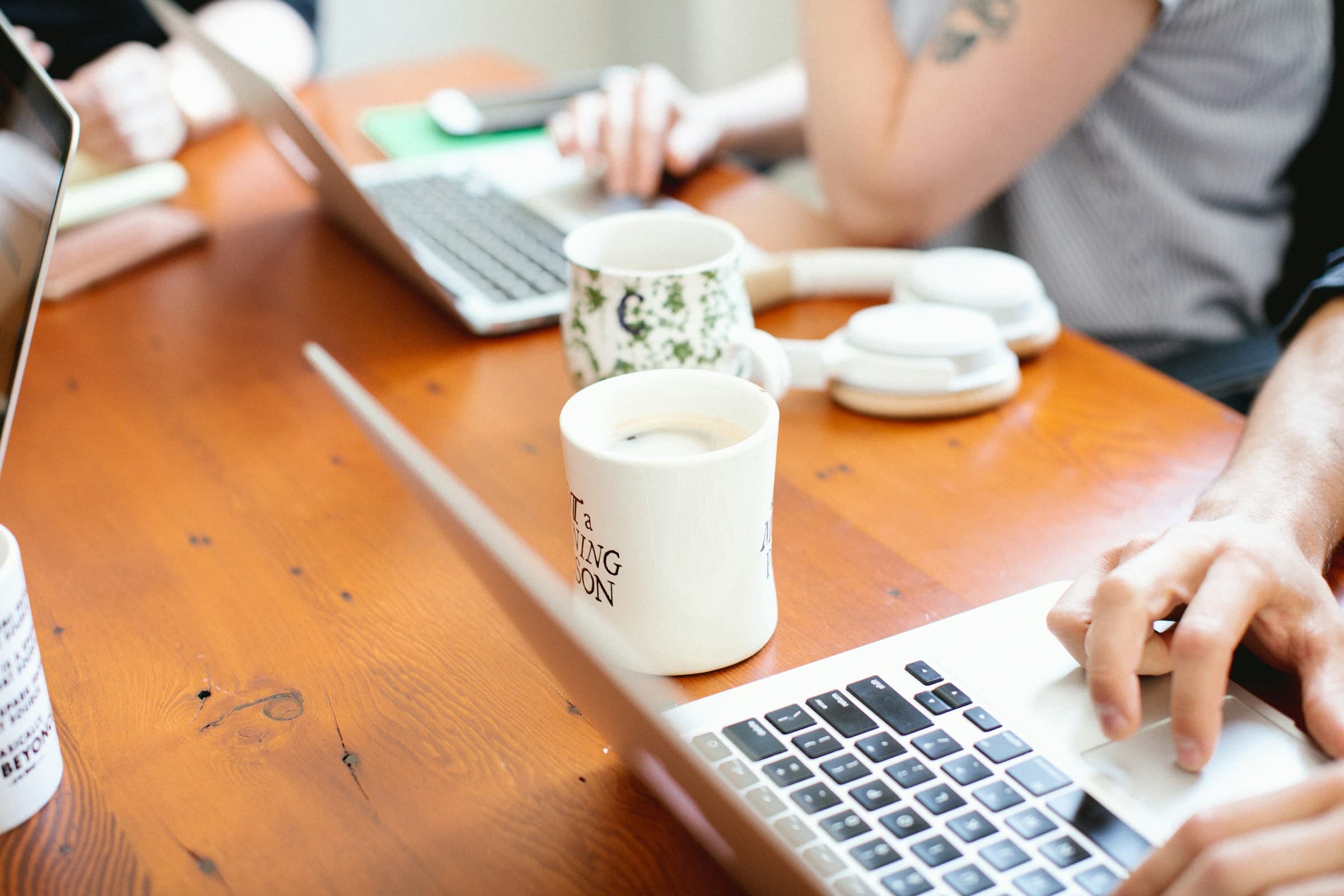 Handen aan een houten tafel met laptops en koffiemokken.