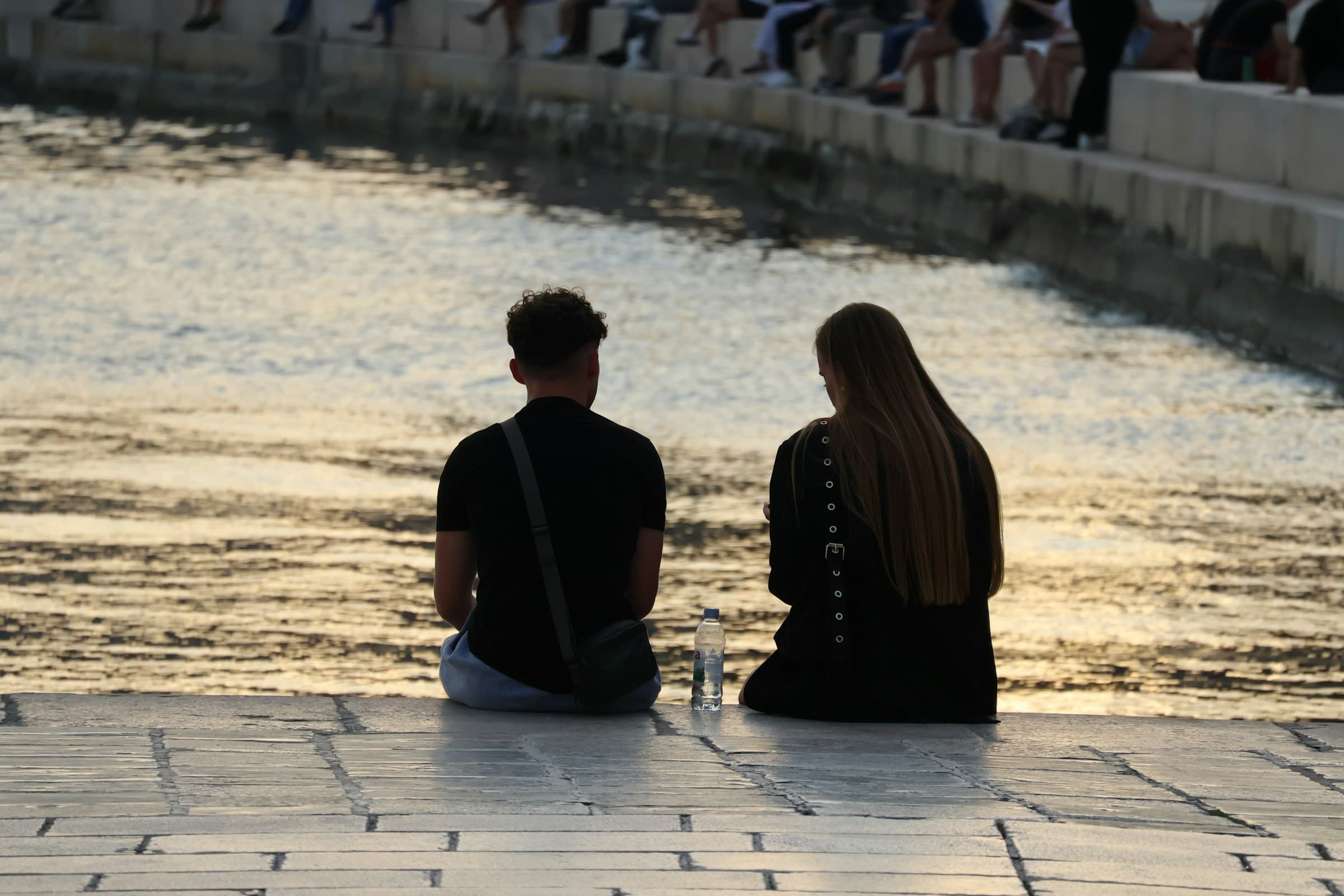 Twee mensen zitten naast elkaar aan het water, van achteren gezien.